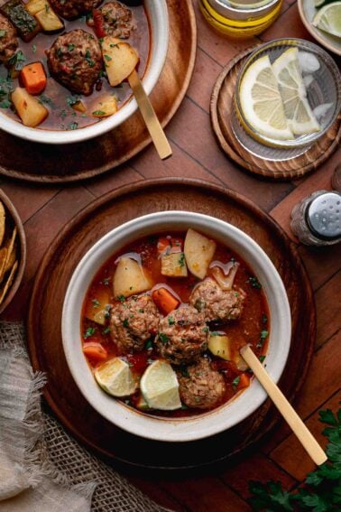 A bowl of albondigas soup with potatoes, carrots, and herbs, served with lemon wedges and a gold spoon on a wooden plate.