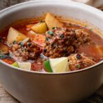 A bowl of albondigas soup with meatballs, potato chunks, and a tomato-based broth, garnished with chopped herbs and lime wedges.