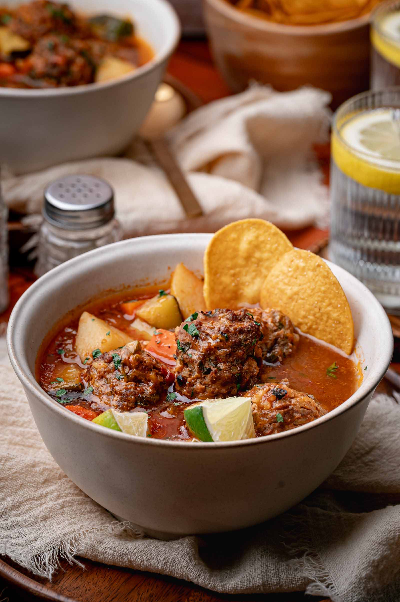 A bowl of Mexican meatball soup with potato chunks, tomato broth, garnished with lime wedges and tortilla chips, sits on a cloth napkin with a drink and spices in the background.