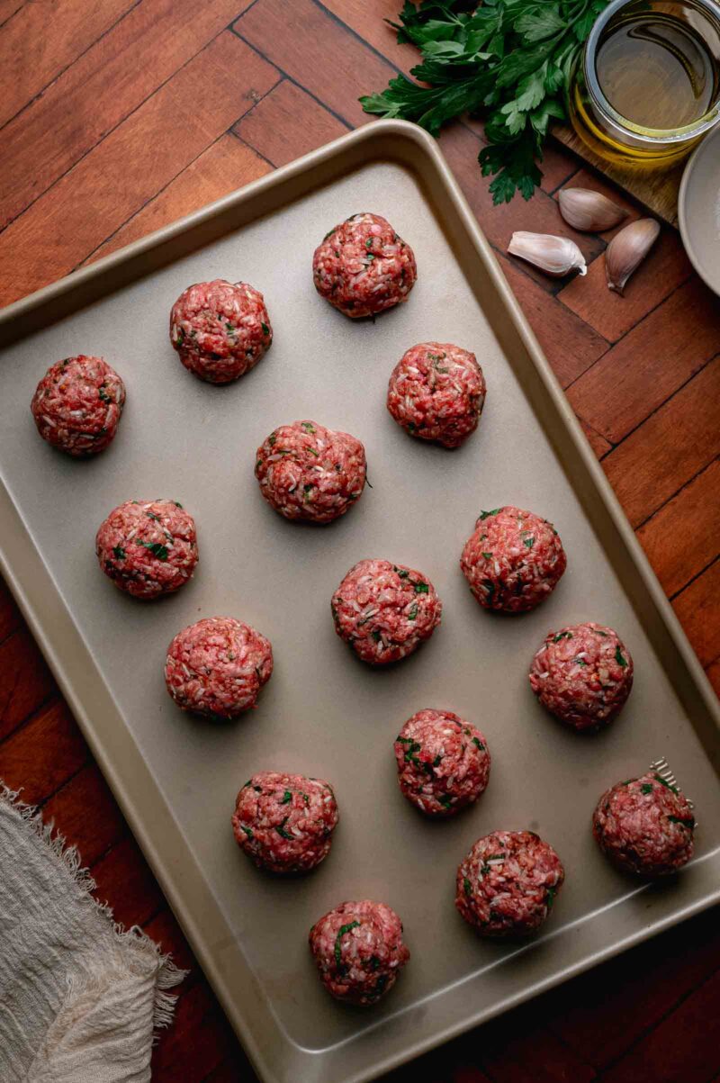 A baking sheet with fifteen raw meatballs spaced evenly, surrounded by parsley, garlic cloves, olive oil, and a white bowl on a wooden surface.