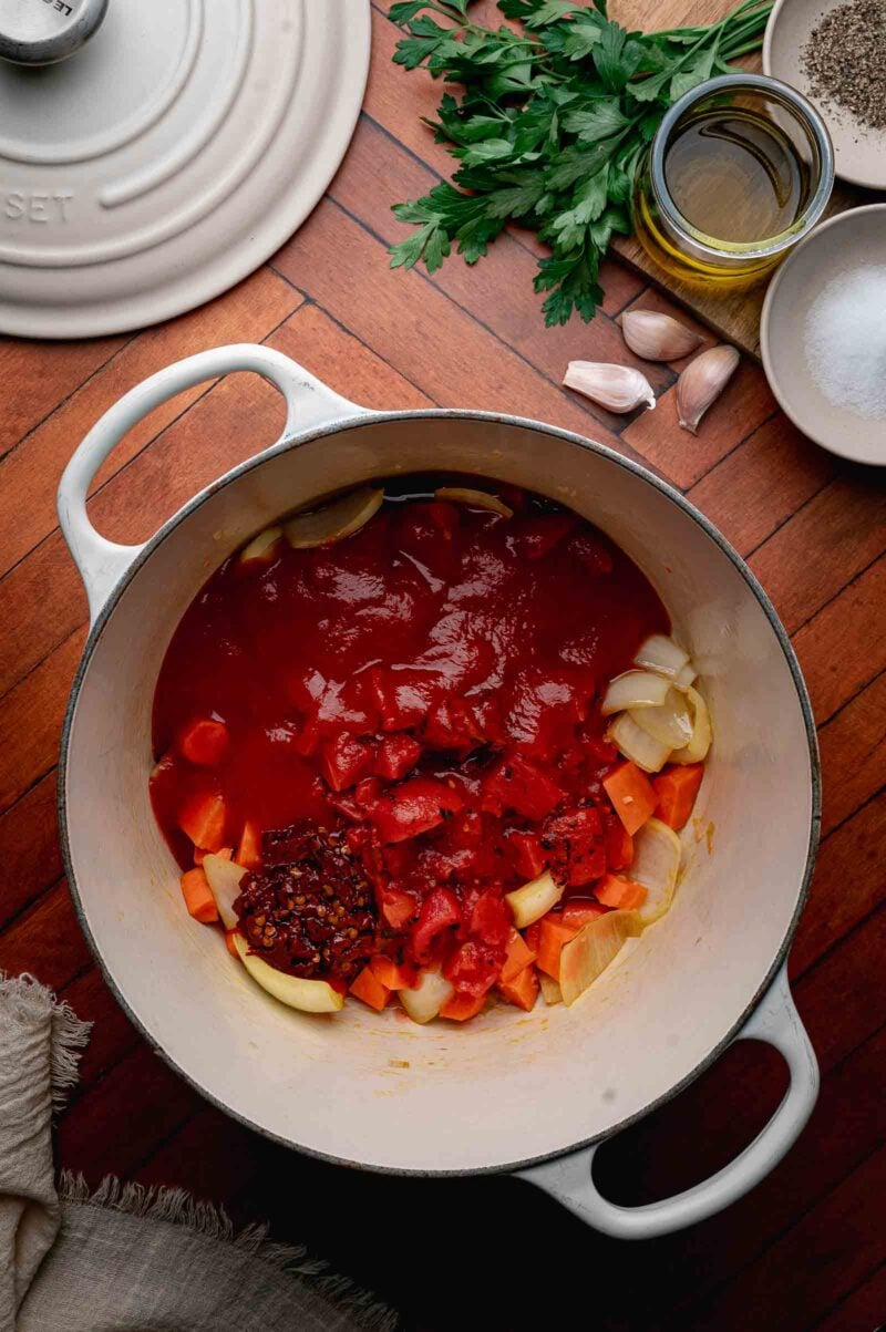 A white Dutch oven on a wooden surface contains chopped onions, carrots, tomatoes, tomato sauce, and chili flakes. Nearby are parsley, garlic, olive oil, salt, and pepper.