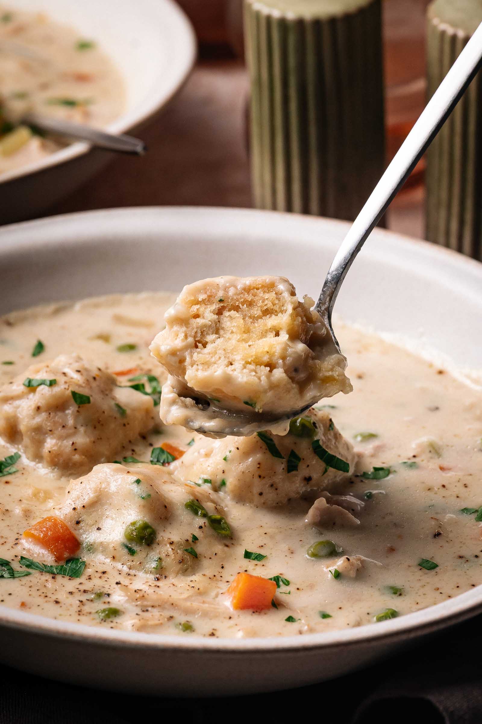 A spoonful of dumpling and vegetables held above a bowl of creamy stew with chicken, carrots, peas, and herbs.
