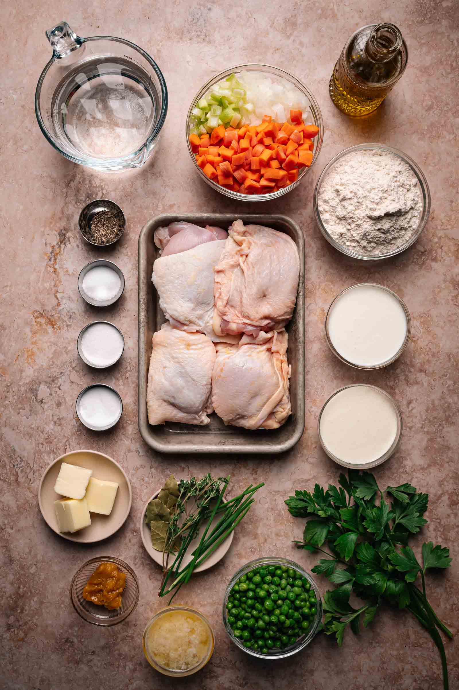 Overhead view of ingredients for chicken pot pie, including raw chicken thighs, chopped vegetables, flour, cream, butter, seasonings, herbs, green peas, and water arranged on a countertop.