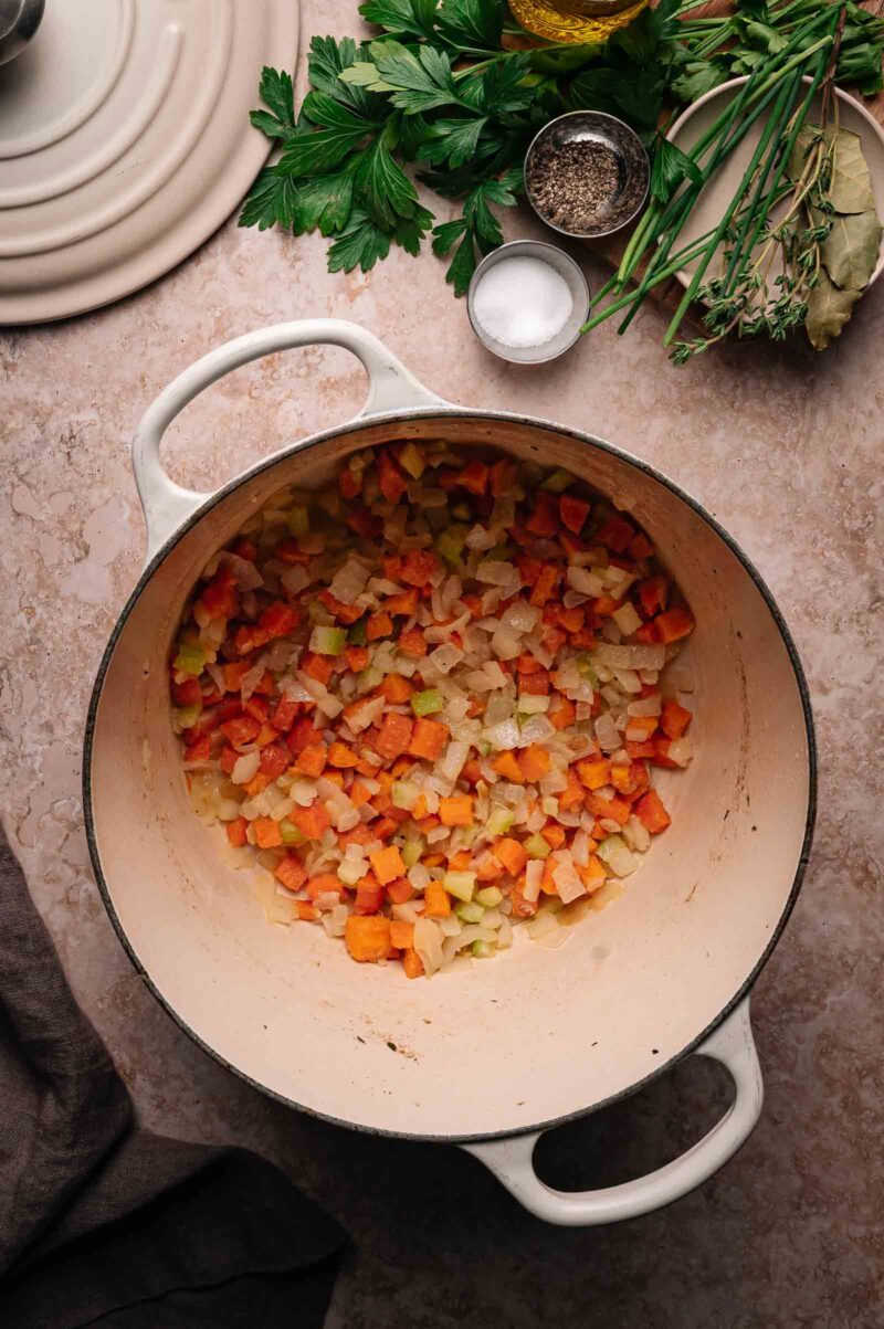 A white dutch oven containing chopped carrots, celery, and onions being sautรฉed; fresh herbs, salt, and pepper are arranged nearby on a marble countertop.