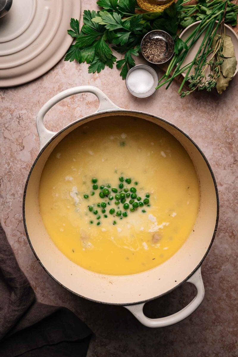 A white pot filled with creamy yellow soup topped with green peas, surrounded by fresh herbs, salt, and pepper on a beige countertop.