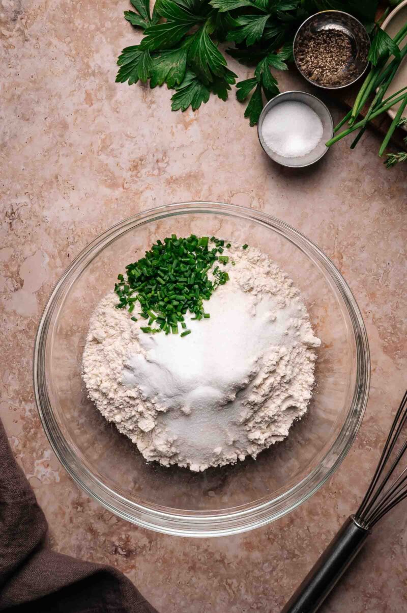 A glass bowl with flour, sugar, and chopped chives on a countertop, surrounded by parsley, salt, pepper, and a metal whisk.
