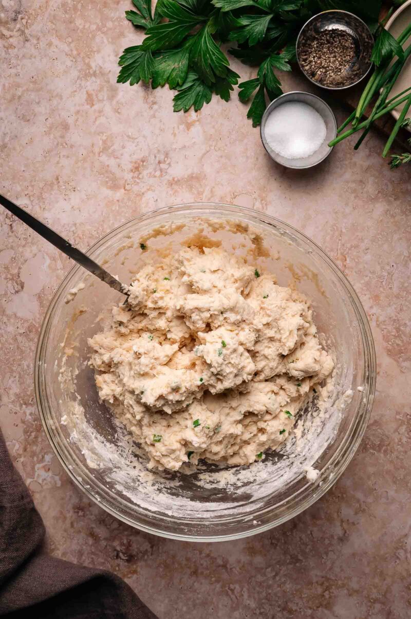 A glass bowl filled with biscuit dough mixed with chopped herbs, a spoon inside, and fresh herbs, salt, and pepper in small bowls nearby on a countertop.