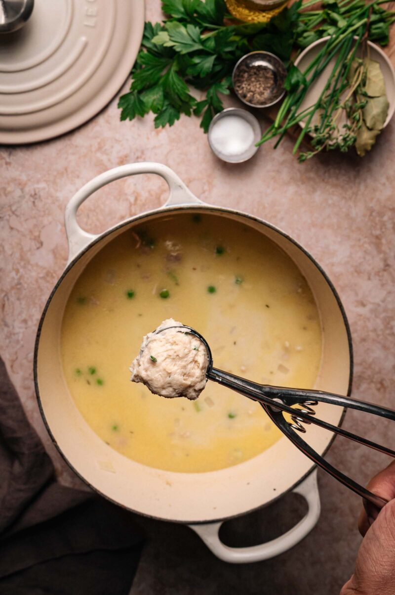 A hand holds a scoop of buttermilk dumplings over a pot of creamy soup with peas, surrounded by herbs and seasonings on a countertop.