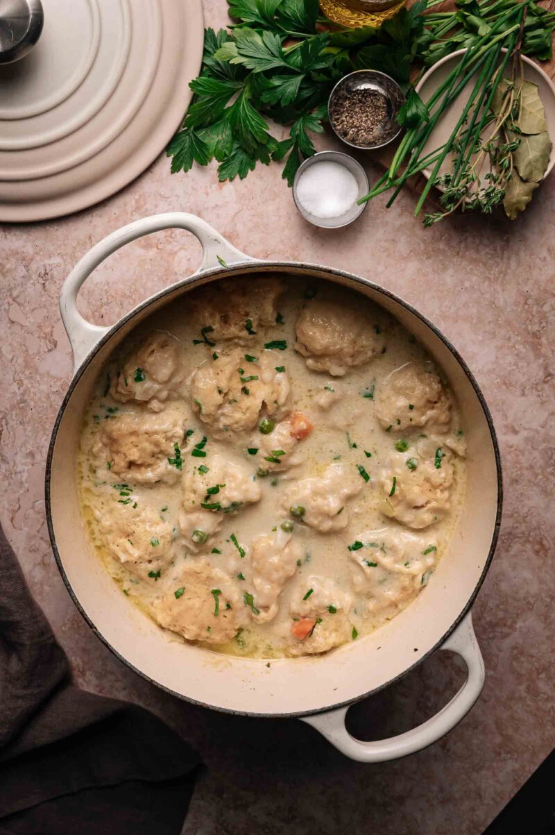 A white pot filled with creamy chicken and dumplings, garnished with chopped herbs, sits on a countertop next to fresh herbs, salt, and pepper.
