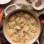 A pot of creamy chicken and dumplings garnished with black pepper and herbs, surrounded by carrots, seasonings, and utensils on a kitchen counter.