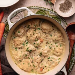 A pot of creamy chicken and dumplings garnished with black pepper and herbs, surrounded by carrots, seasonings, and utensils on a kitchen counter.