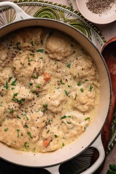 A pot of creamy chicken and dumplings garnished with chopped parsley, with black pepper sprinkled on top, sits on a woven placemat next to a small dish of ground pepper.