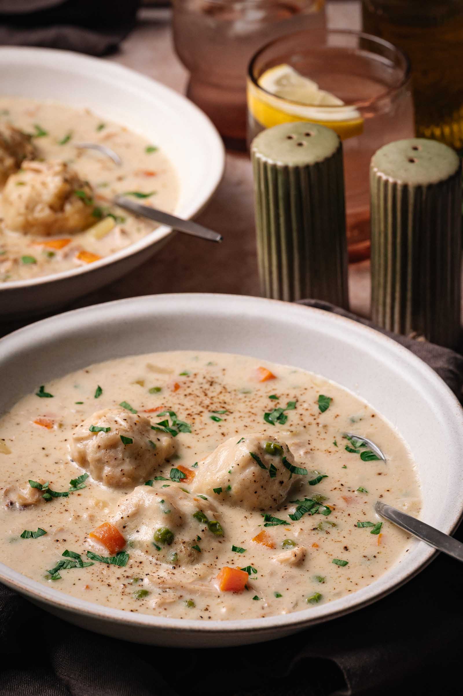 A bowl of creamy chicken and dumplings, garnished with herbs, sits next to a spoon and green salt and pepper shakers.