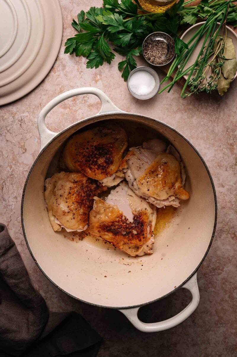 Four seasoned chicken thighs are browning in a white Dutch oven, with fresh herbs, salt, pepper, and olive oil arranged nearby on a beige countertop.