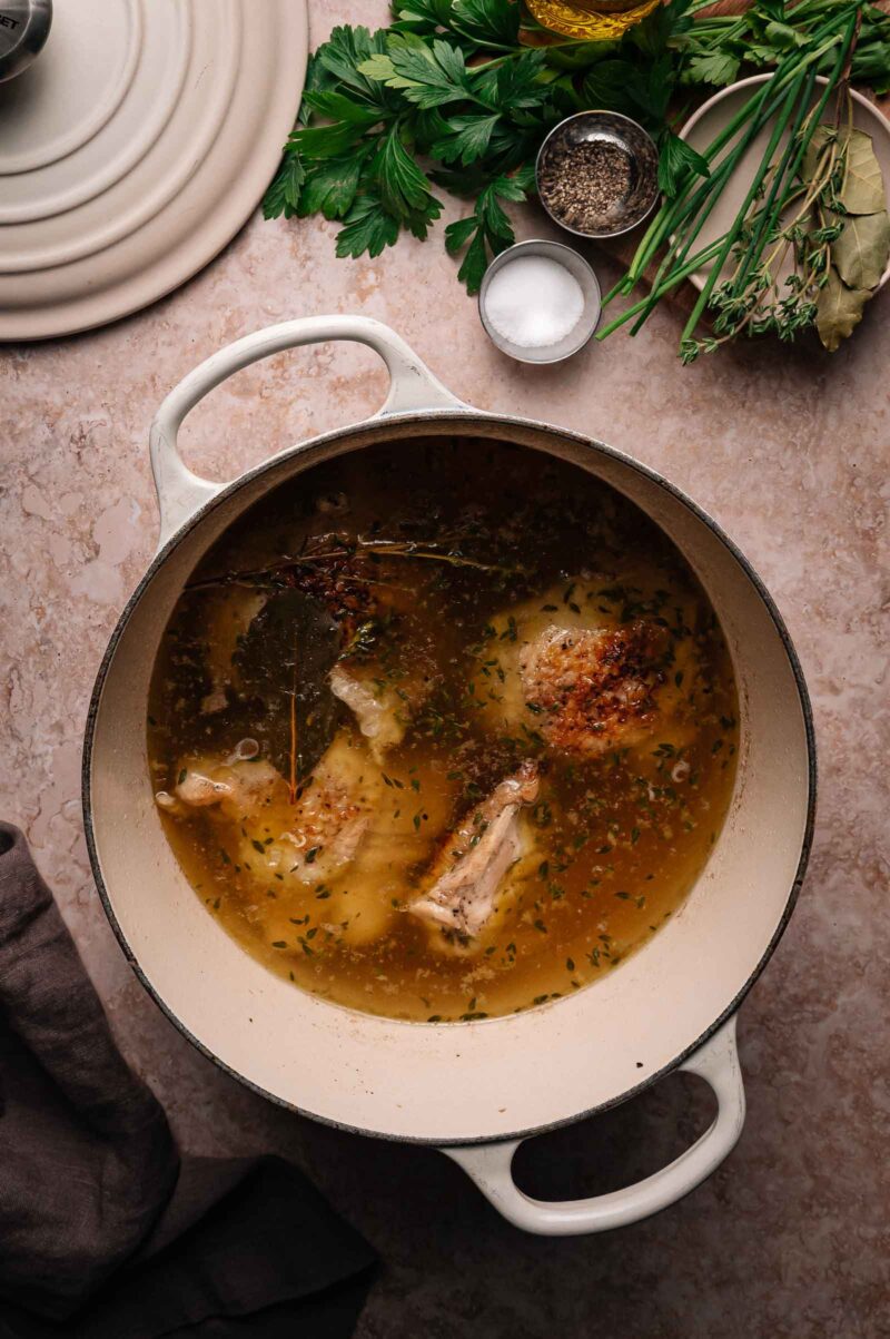 A white Dutch oven filled with simmering chicken broth and herbs sits on a countertop next to fresh herbs, salt, and pepper.