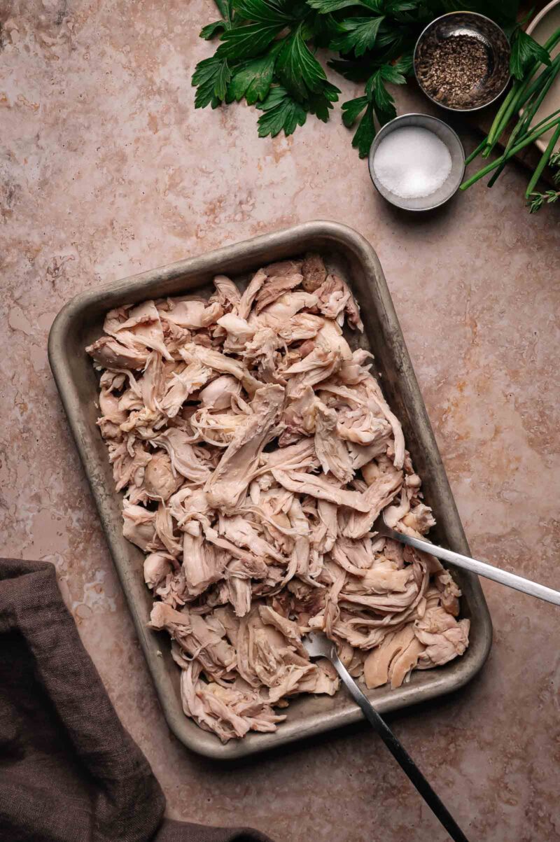 A baking tray filled with shredded cooked chicken, with two forks for pulling the meat apart. Fresh herbs, salt, and pepper are placed nearby on a marble countertop.
