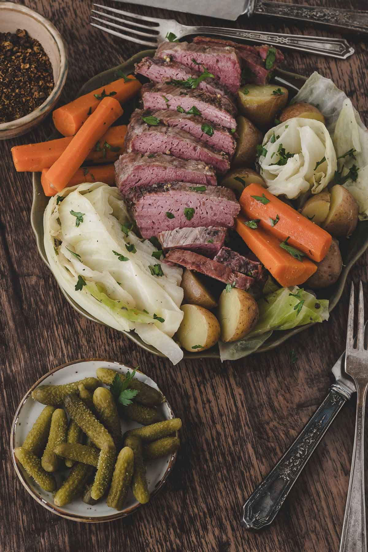 Sliced corned beef served with boiled potatoes, carrots, and cabbage on a plate, with a side dish of pickles and utensils on a wooden table.