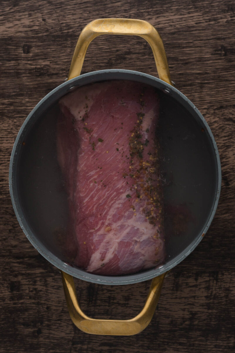 A raw corned beef brisket sits in a pot filled with water and spices on a wooden surface, viewed from above.