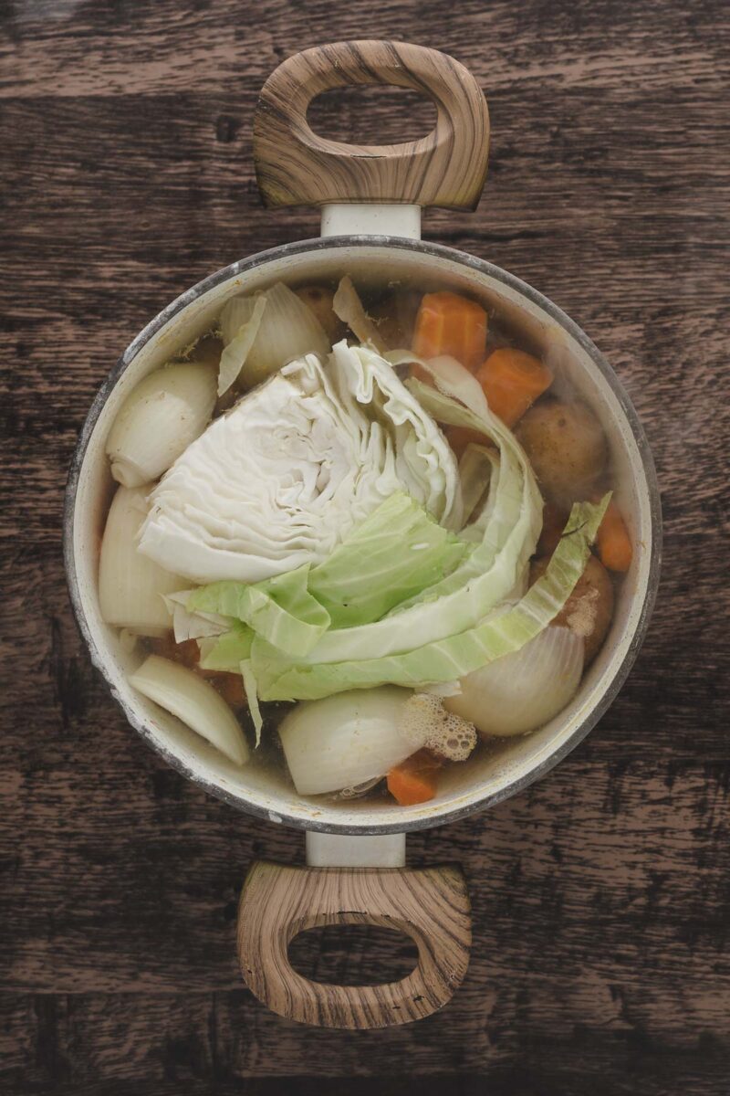 A pot of soup containing cabbage, onions, carrots, and potatoes sits on a wooden surface, viewed from above.