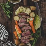 Sliced corned beef with carrots, potatoes, and cabbage on a wooden board, garnished with parsley; pickles and a bowl of spices are beside the board.