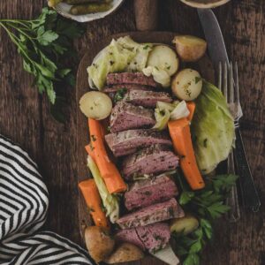 Sliced corned beef with carrots, potatoes, and cabbage on a wooden board, garnished with parsley; pickles and a bowl of spices are beside the board.