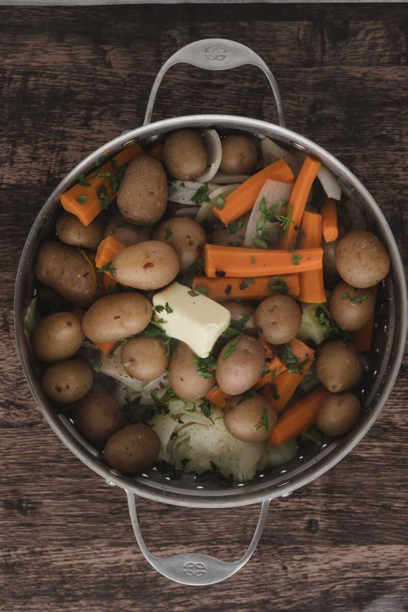 Overhead view of a pot filled with small potatoes, carrot sticks, chopped onions, fresh herbs, and two pats of butter, placed on a wooden surface.