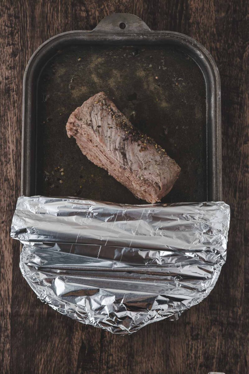 A cooked beef roast sits in a dark roasting pan on a wooden surface, partially covered with crumpled aluminum foil.