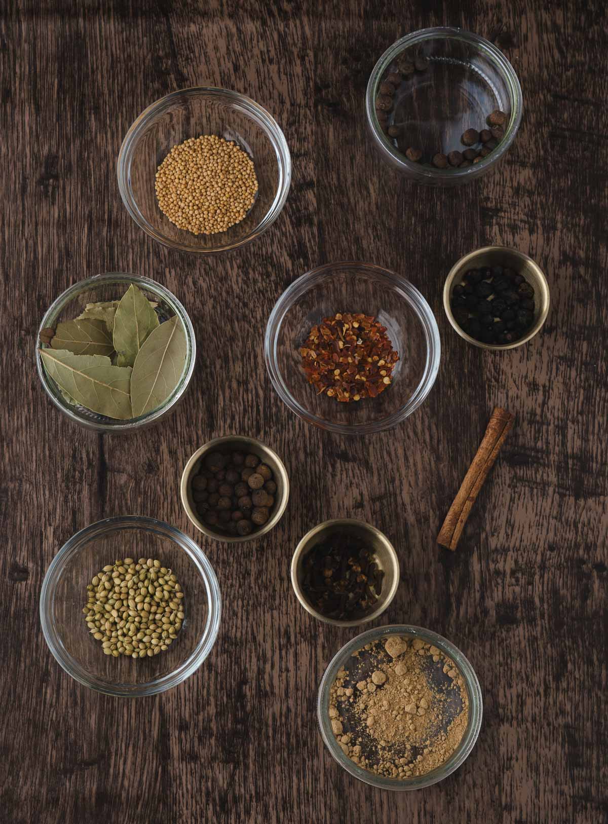 Nine glass and ceramic bowls filled with various whole and ground spices, plus a cinnamon stick, arranged on a dark wooden surface.