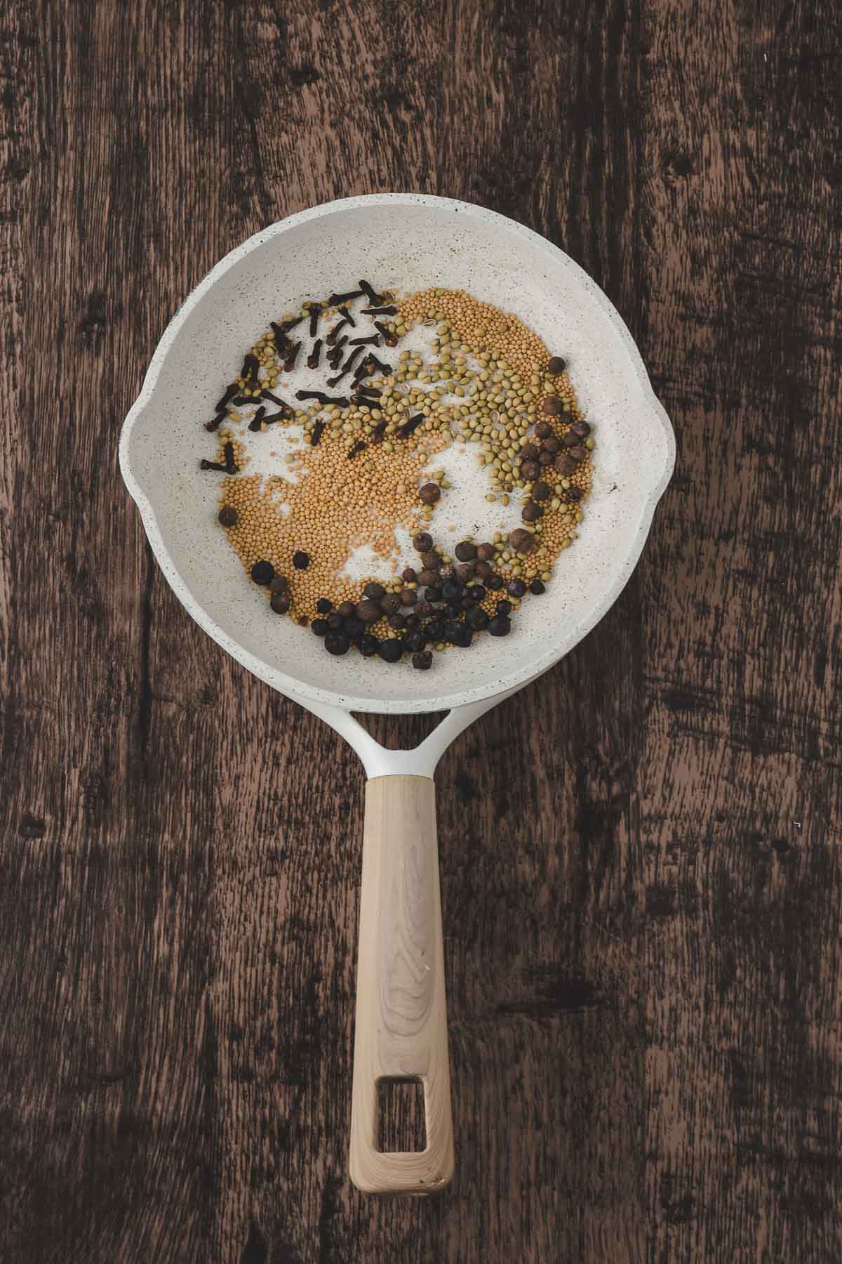 A white frying pan with whole spices, including mustard seeds, coriander seeds, cloves, and black peppercorns, on a dark wooden surface.