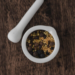 A white mortar and pestle containing a mix of whole spices, including mustard seeds and peppercorns, sits on a dark wooden surface.