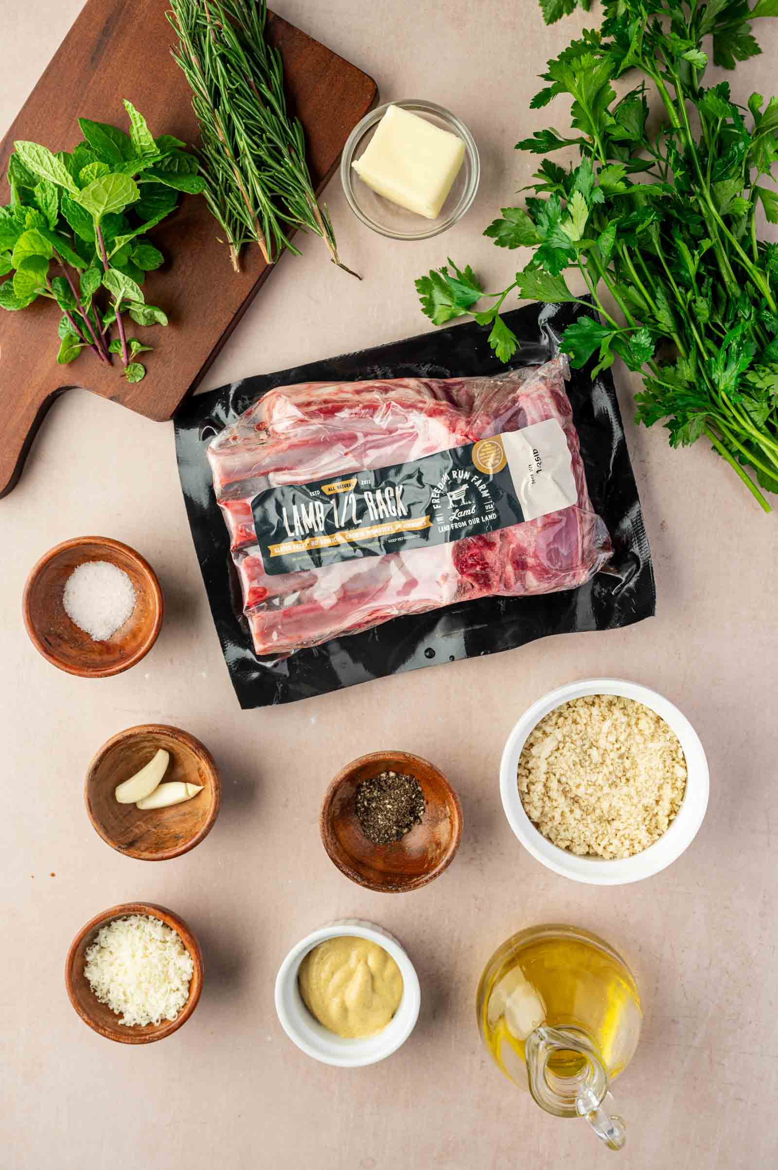 Ingredients for lamb dish arranged on a countertop, including lamb rack in packaging, herbs, butter, salt, garlic, cheese, mustard, breadcrumbs, oil, and pepper.