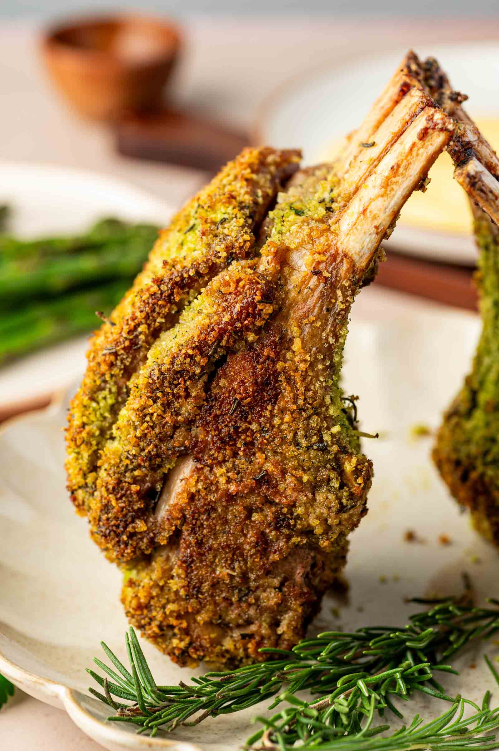 A rack of lamb coated in a golden herb and breadcrumb crust, standing upright on a plate with sprigs of rosemary and asparagus in the background.