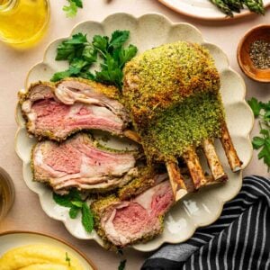 A sliced rack of herb-crusted lamb is served on a plate with parsley, alongside asparagus, polenta, olive oil, and a striped napkin.