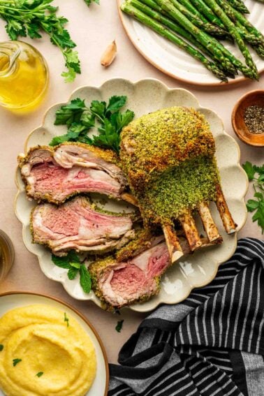 A sliced rack of herb-crusted lamb is served on a plate with parsley, alongside asparagus, polenta, olive oil, and a striped napkin.