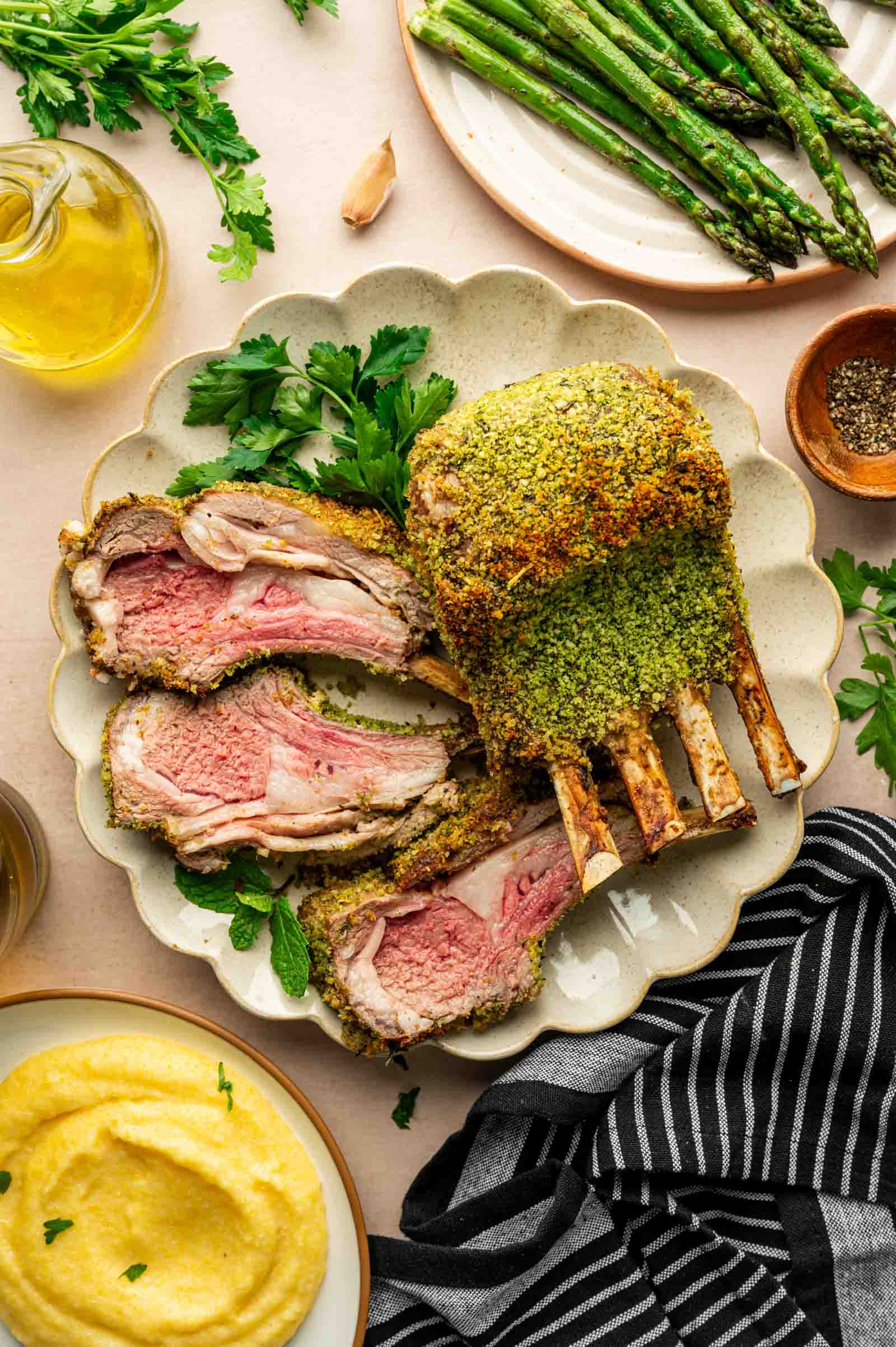 A sliced rack of herb-crusted lamb is served on a plate with parsley, alongside asparagus, polenta, olive oil, and a striped napkin.