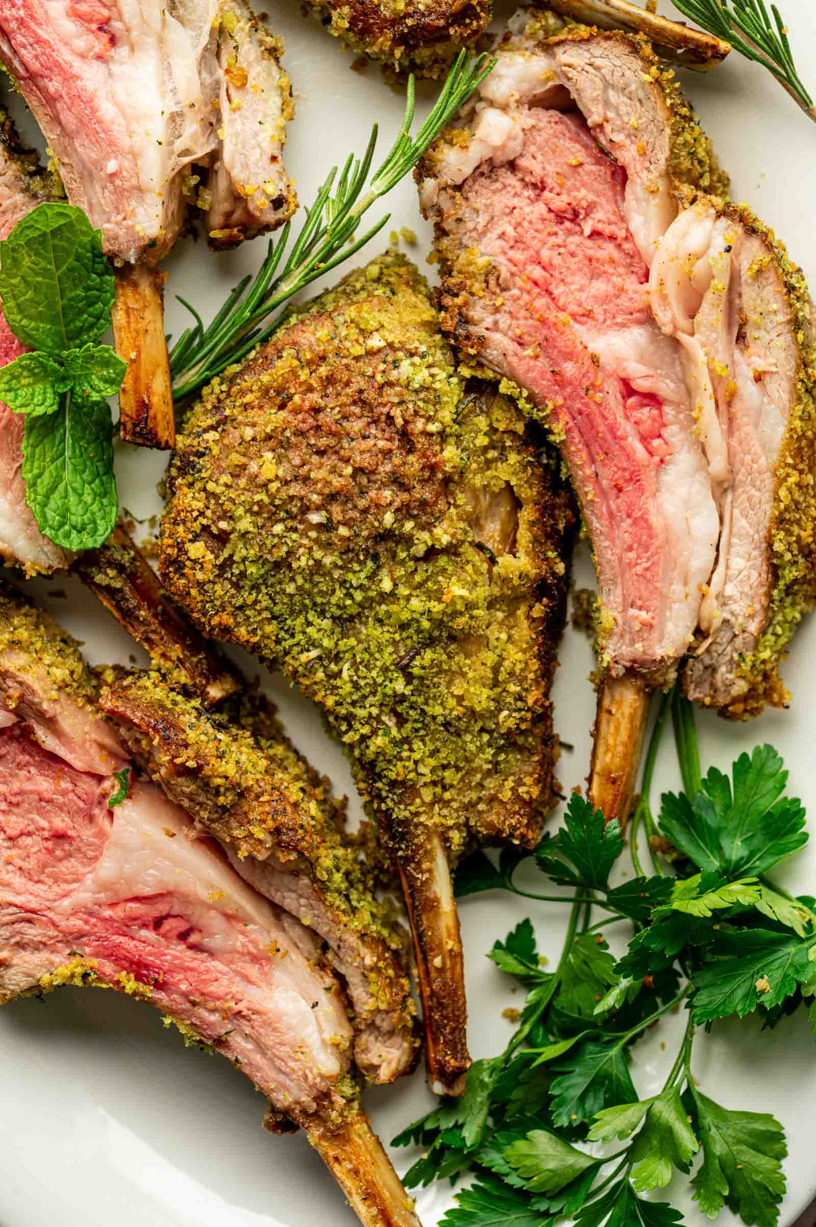 Close-up of several slices of herb-crusted rack of lamb, cooked medium-rare, garnished with fresh rosemary, mint, and parsley on a white plate.