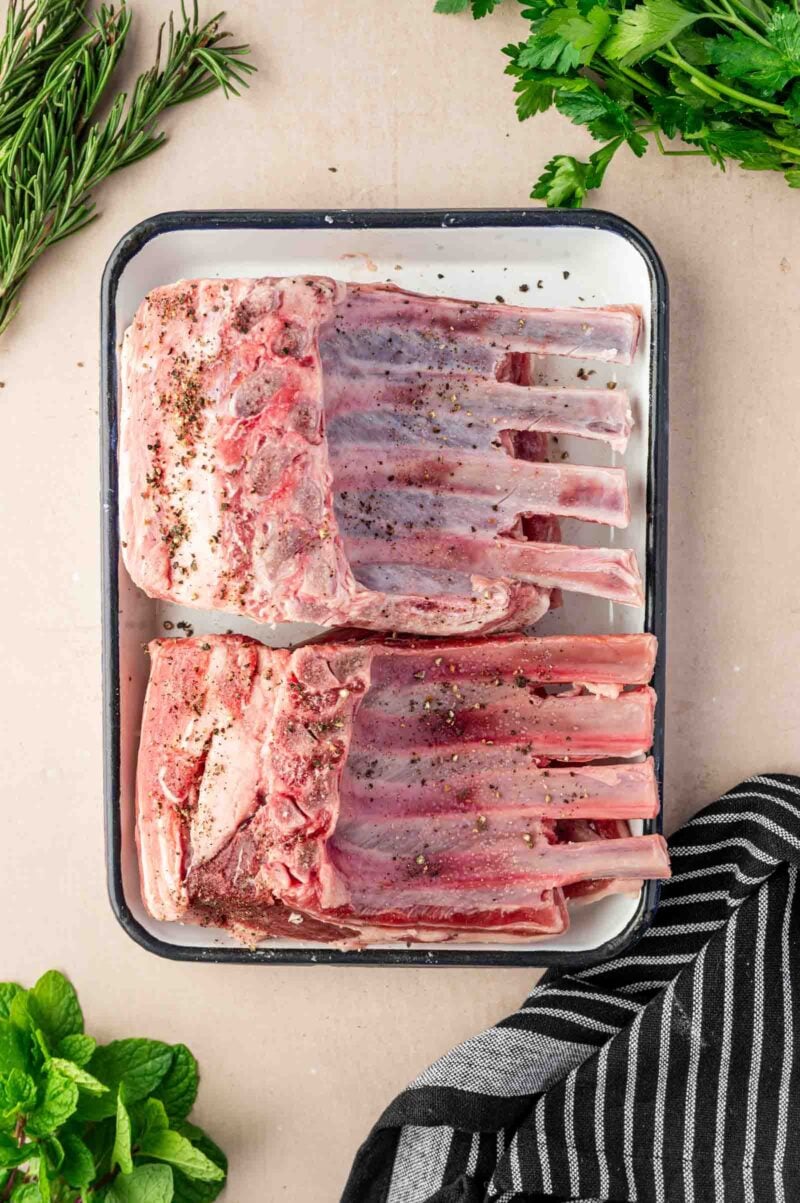 Two raw racks of lamb seasoned with salt and pepper on a white tray, surrounded by fresh herbs and a striped cloth.