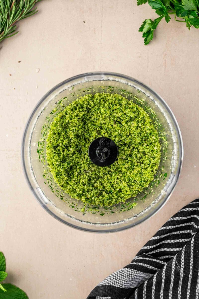 Overhead view of green herb mixture blended in a food processor, with fresh herbs and a striped cloth visible nearby.