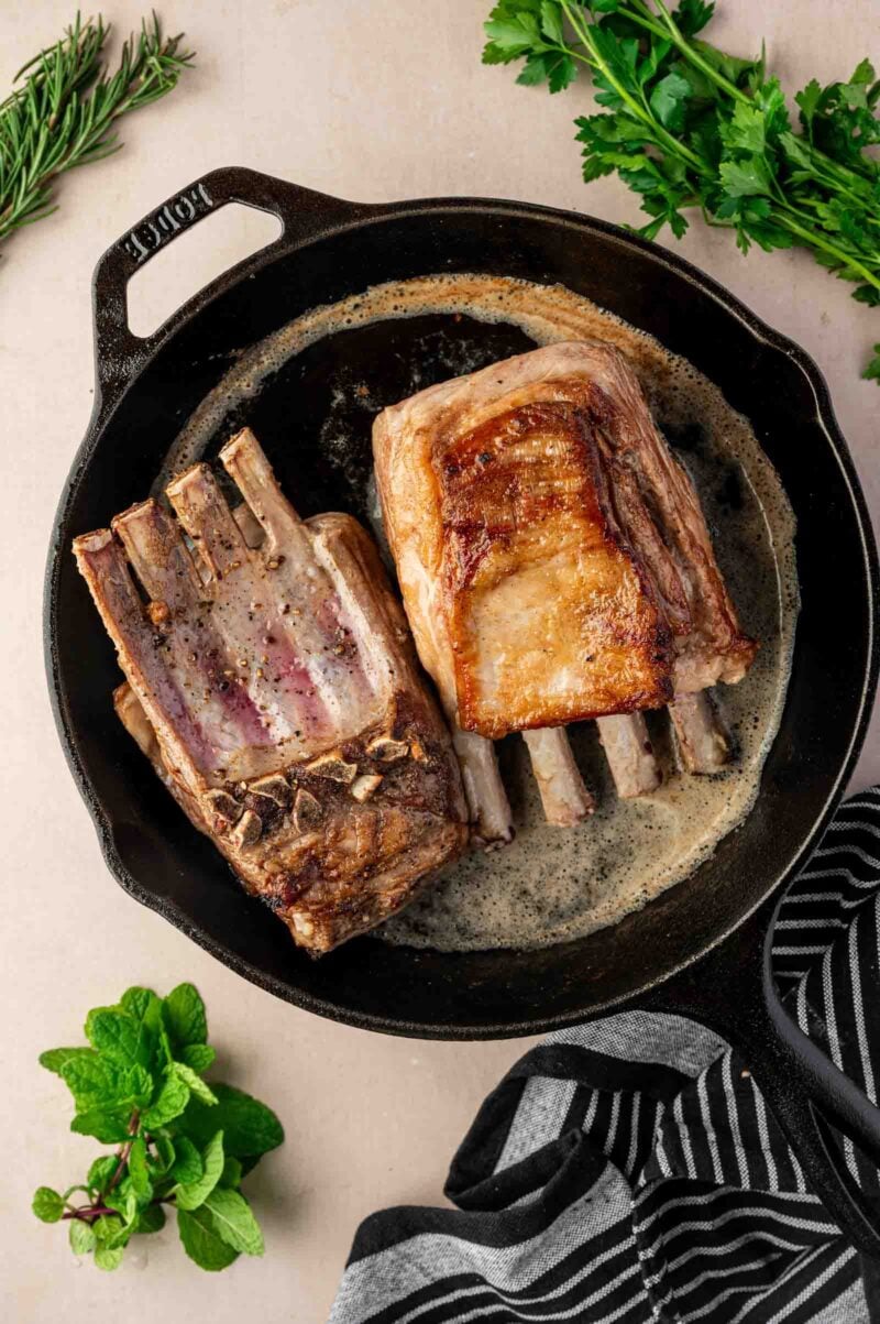 Two racks of lamb are searing in a cast iron skillet with bubbling fat, surrounded by fresh herbs on a light countertop.