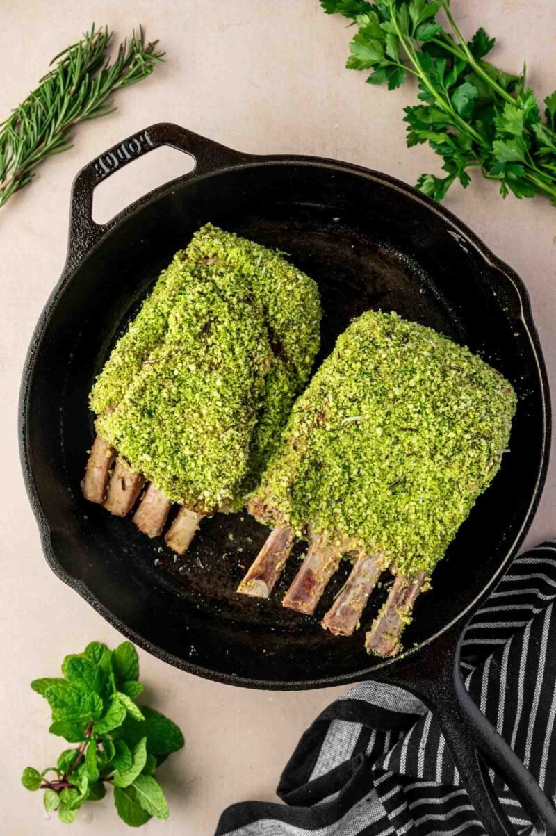 Two herb-crusted racks of lamb in a black cast iron skillet, surrounded by fresh herbs on a light surface.