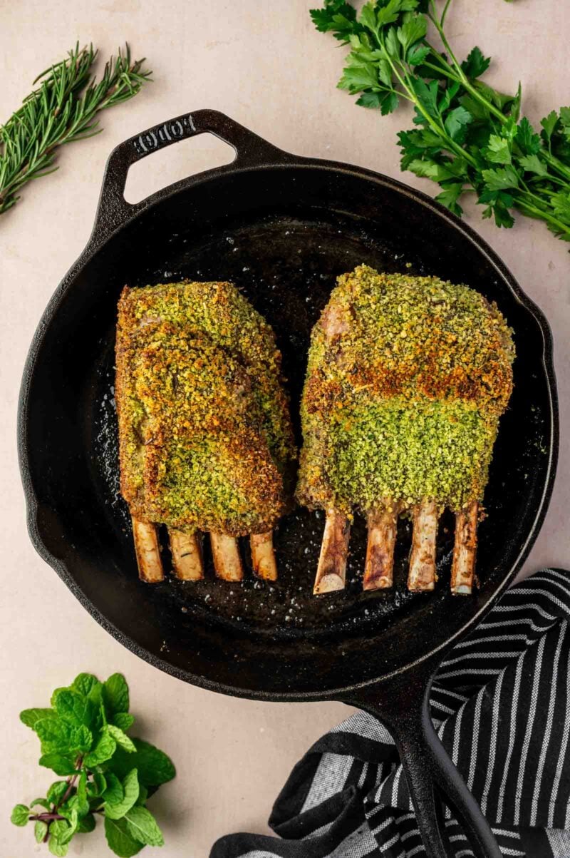 Two herb-crusted racks of lamb in a black cast iron skillet, surrounded by fresh rosemary, parsley, mint, and a striped kitchen towel.