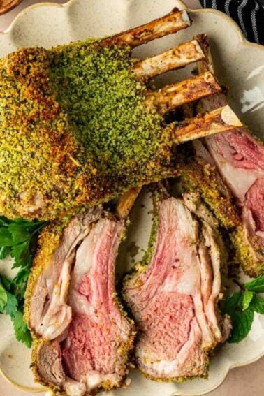 A rack of lamb with a green herb crust is sliced and served on a plate with fresh herbs, alongside asparagus and polenta.