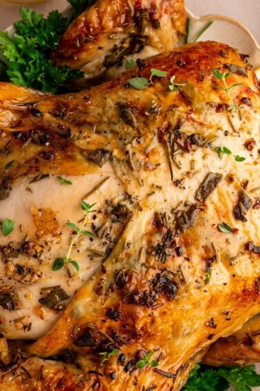 A whole roasted chicken garnished with herbs is served on a plate, surrounded by fresh parsley and lemon wedges.