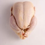 A whole raw chicken with legs tied together, placed on a plain white surface, viewed from above.