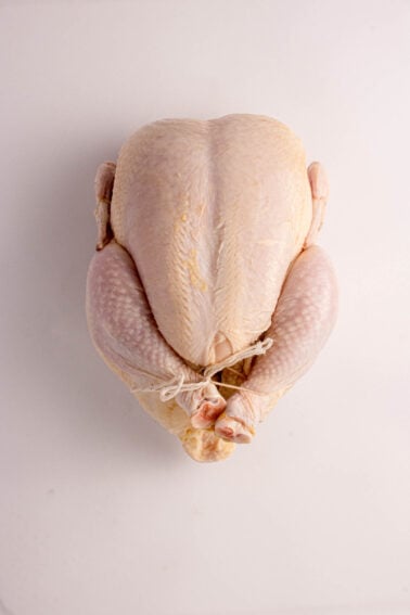 A whole raw chicken with legs tied together, placed on a plain white surface, viewed from above.