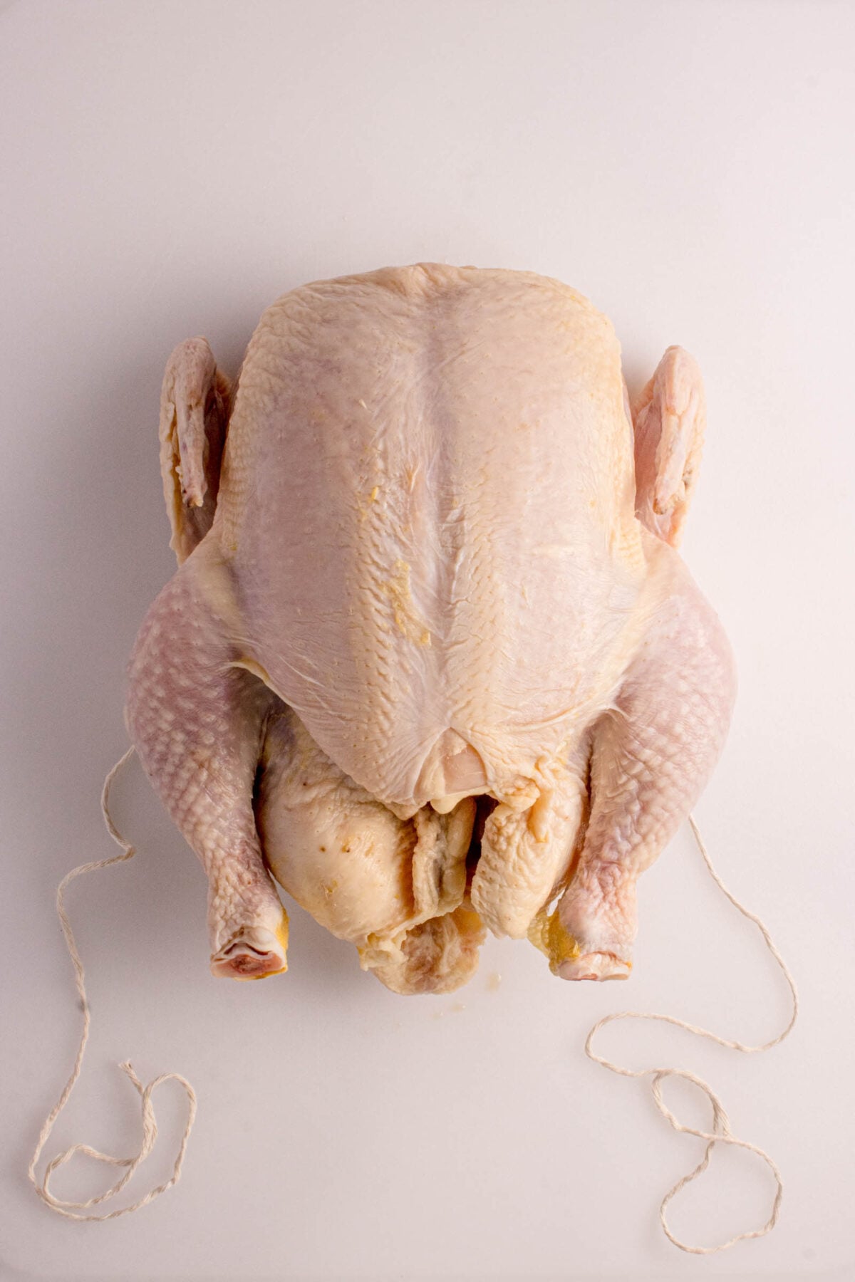 Raw whole chicken with legs tied, placed on a white surface, viewed from above.