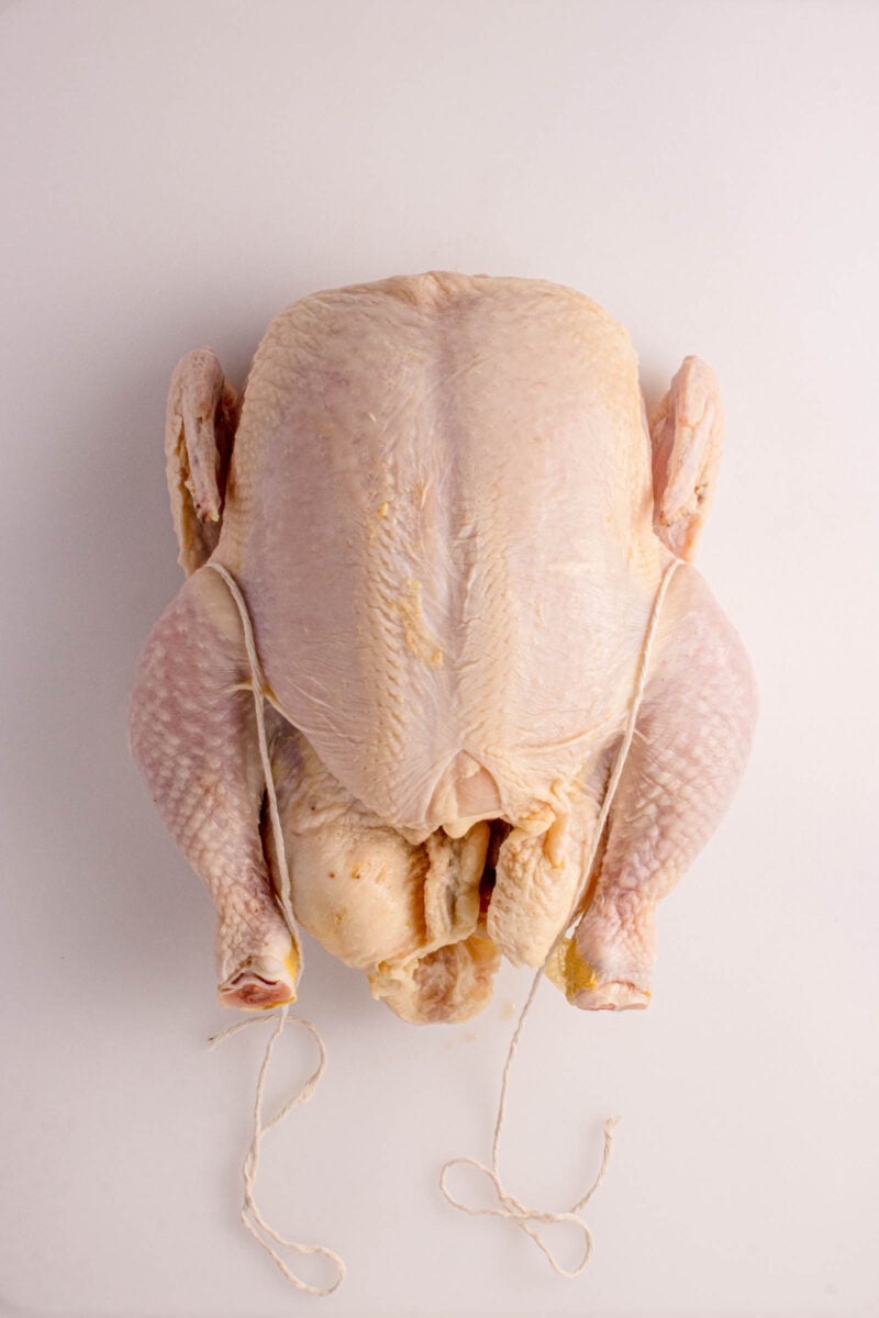Raw whole chicken, trussed with kitchen twine, placed on a plain white surface.