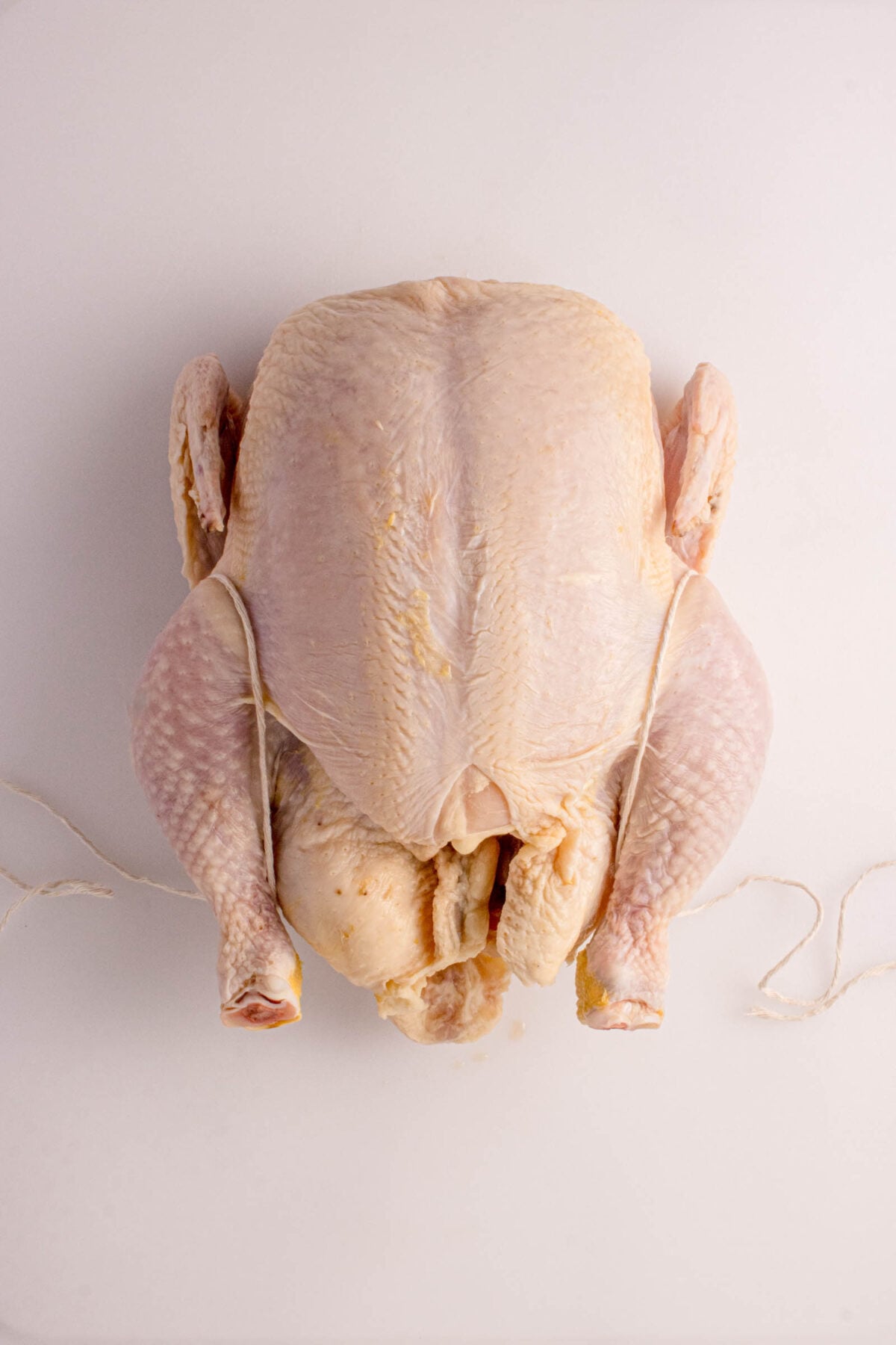 A whole raw chicken with its legs tied together, placed on a white surface.