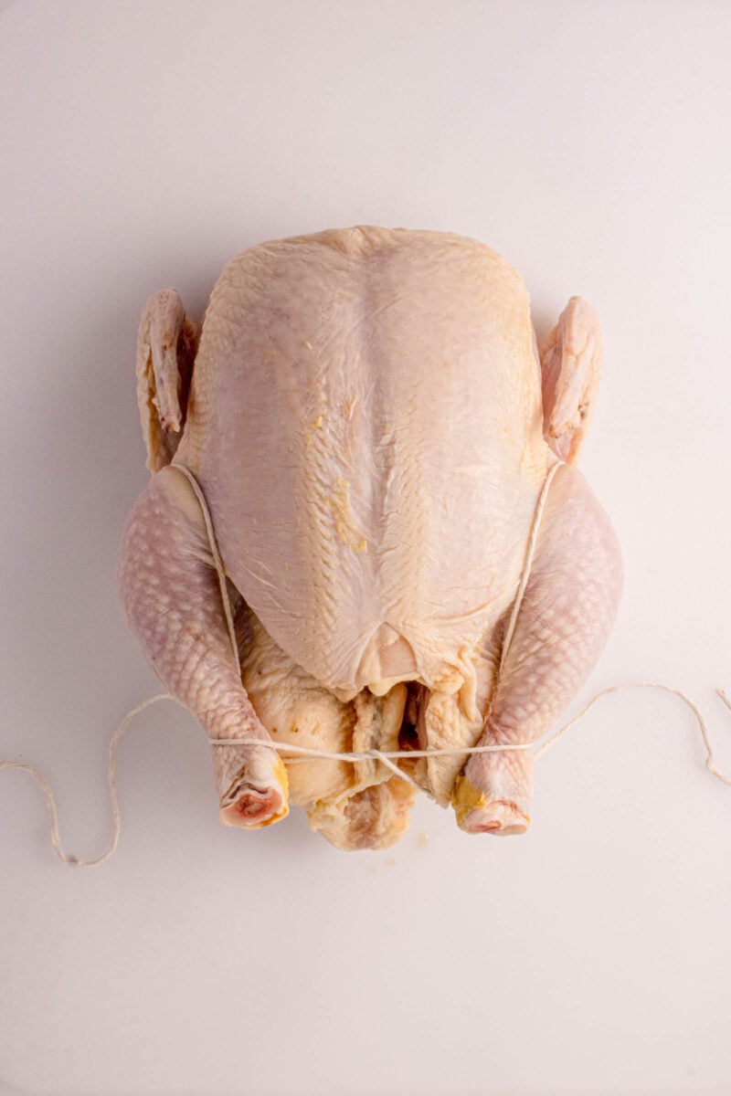 Whole uncooked chicken with legs tied together, placed on a white surface.