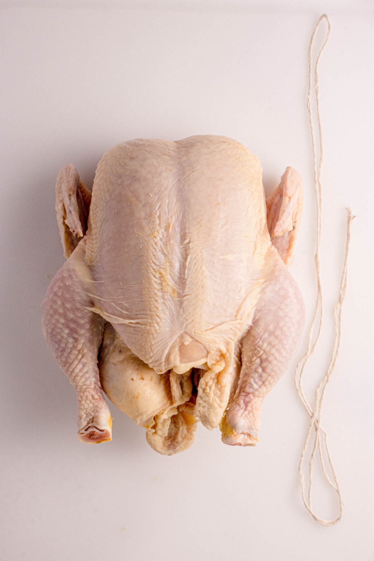 Raw whole chicken placed on a white surface next to a piece of kitchen twine.